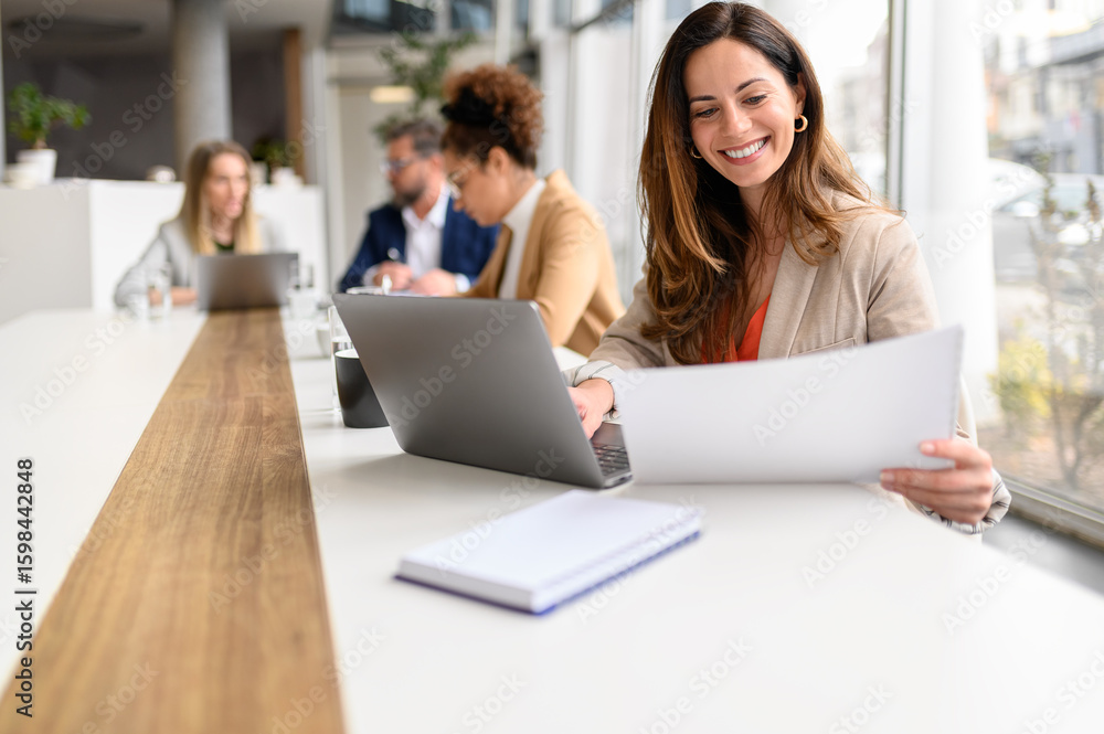 Fototapeta premium Smiling professional analyzing legal document while working on laptop in modern office with coworkers in the background