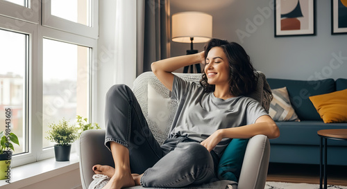 Young relaxed smiling pretty woman relaxing sitting on chair at home. Happy positive beautiful lady feeling joy enjoying wellbeing and lounge chilling near window in modern cozy apartment interior