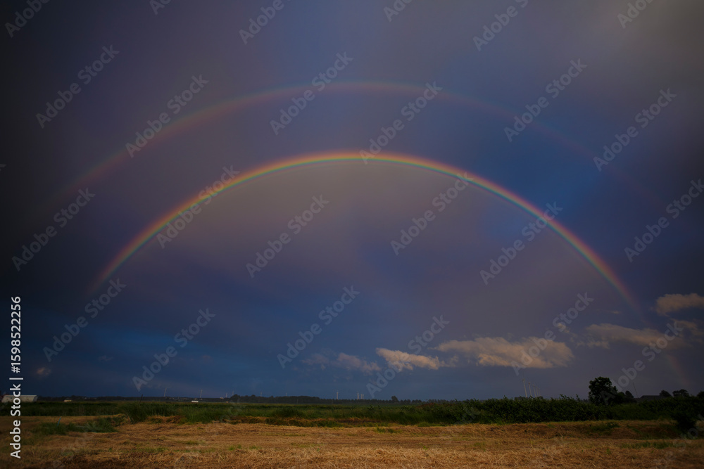 Naklejka premium Colorful rainbow arches over a lush field during a dramatic sky at sunset after a rainstorm in a rural landscape