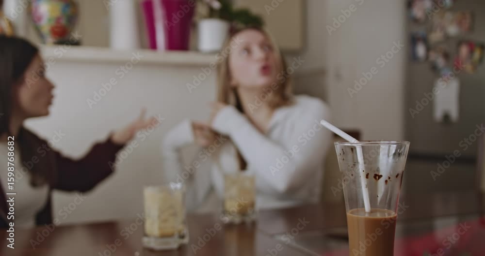 Two female friends enjoy iced coffee together and engage in an animated discussion at home. Authentic moment of connection and friendship. Two female friends enjoy iced coffee together and engage in an animated discussion at home. Authentic moment of connection and friendship.
