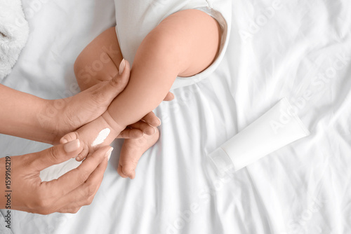 Female hands applying cream onto little baby's foot on bed, top view