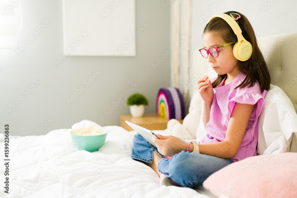 Girl wearing headphones using tablet and eating chips in bed Girl wearing headphones using tablet and eating chips in bed