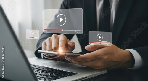 A man using a smartphone with video streaming icons and a laptop in the foreground at a desk