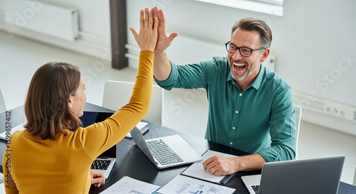 Two colleagues, a man and a woman, celebrate a successful collaboration with a high five in a bright office setting.