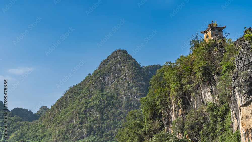 Fototapeta premium Steep mountains against the blue sky. Green forest on the slopes. At the top, you can see a Chinese pagoda with a roof with curved cornice edges. China. Guilin
