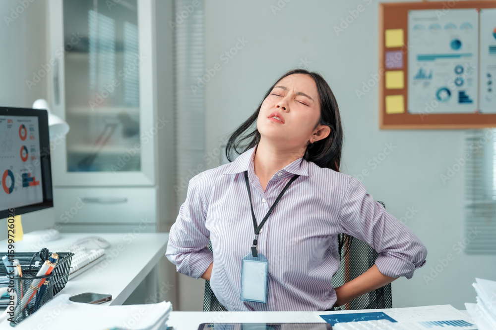 Naklejka premium Young businesswoman gripping her lower back, displaying a pained expression while enduring backache from long hours of sedentary office work, highlighting the toll of an unhealthy lifestyle