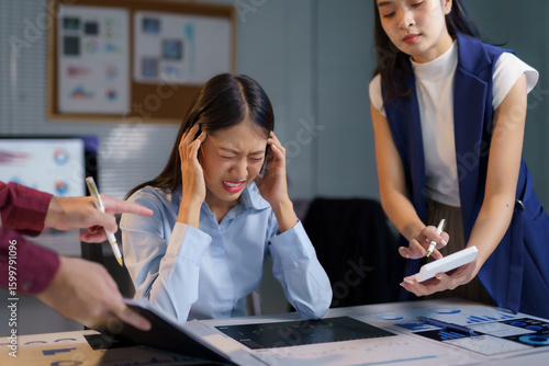 Stressed businesswoman experiencing a headache while working on a financial report, surrounded by colleagues pointing out errors in a hectic office environment