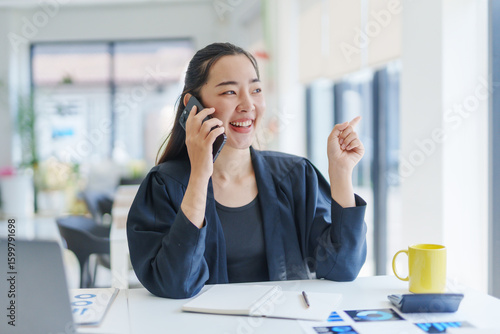 Young happy businesswoman talking on mobile phone and pointing with finger while sitting at desk with laptop, notebook, pen, calculator, charts and mug in office