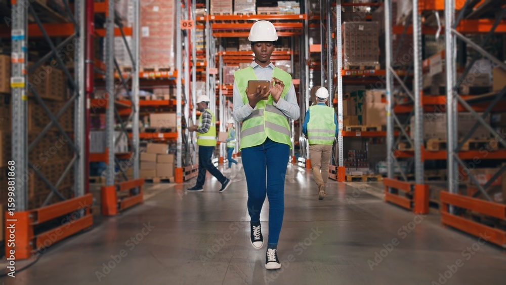 Obraz premium African American young professional woman worker in hard hat uses digital tablet checks inventory walks in retail warehouse full of shelves with goods People working Distribution Center Following shot