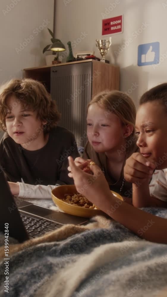 Vertical shot of diverse young friends lying on bed watching movie on laptop while enjoying sweet popcorn together during summer vacation in cozy bedroom Vertical shot of diverse young friends lying on bed watching movie on laptop while enjoying sweet popcorn together during summer vacation in cozy bedroom