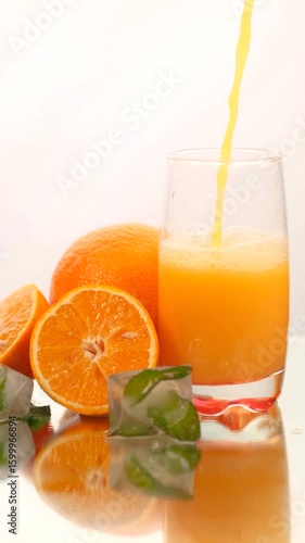 Oranges, a glass and some ices in an image composition on a white background, while they pour orange juice into the glass and the shot opens slowly.
