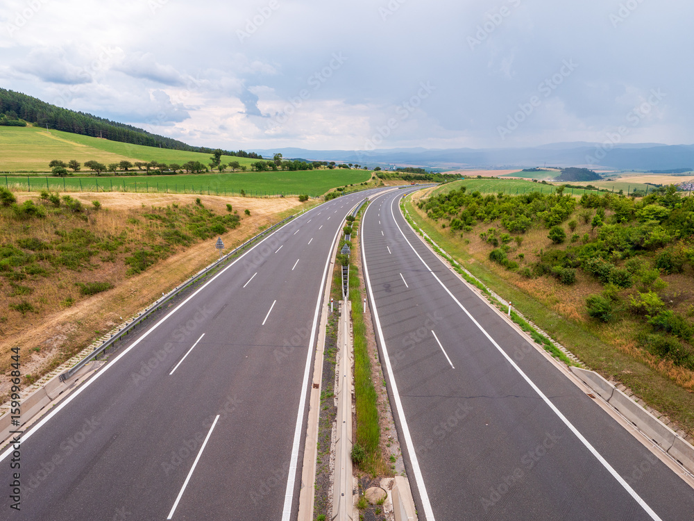 Fototapeta premium Straight empty D1 highway in rural Slovakia near Spišský Hrhov. Green fields, hills and cloudy sky in summer landscape.