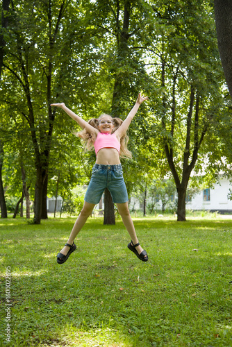 funny happy smiling little girl in glasses in pink top and jeans  jumping in park on sunny summer day