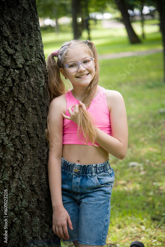 funny happy smiling little girl in glasses in pink top and jeans  posing in park on sunny summer day