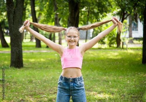 funny happy smiling little girl in glasses in pink top and jeans  posing in park on sunny summer day