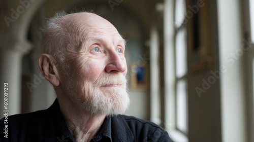 Elderly man with white beard and thoughtful expression looking out window indoors.