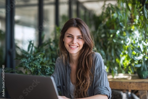 Free Download Happy young woman smiling using laptop in lush greenhouse with green plants and sunlight. - GFXLoad