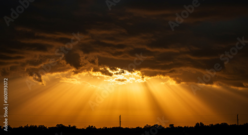 Dramatic god rays and golden sunbeams bursting through dark storm clouds at sunset. A spiritual, religious background symbolizing hope, heaven, and faith over a landscape silhouette. Epic sky.
