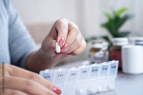 close-up view of a woman organizing pills in a plastic pill box at home, preparing daily medication and vitamins for the week, showing a healthcare routine and medicine management