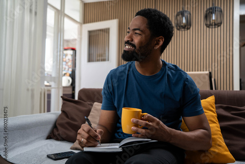 Thoughtful handsome Black man looking away smiling in living room, happy male reflecting on ideas and positive thoughts