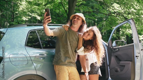 A young couple takes a selfie on their phone against the background of a car and a summer forest, preserving memories of the trip