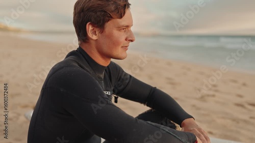 A surfer in a wetsuit sits on his surfboard on the beach, watching the waves and waiting for the right moment to go in.