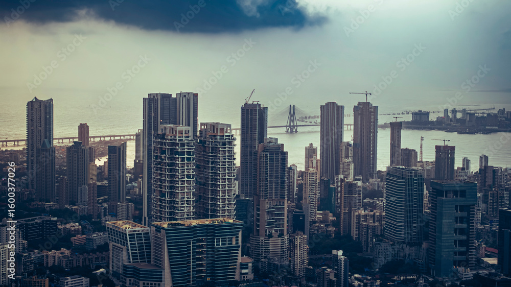 Obraz premium Aerial view of Mumbai city during a monsoon evening with dramatic, cloudy skies. Bandra-Worli Sea Link is visible behind high-rise buildings, a scenic urban and weather contrast.
