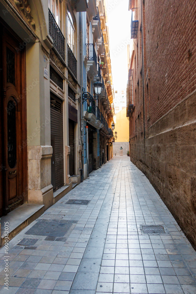 Obraz premium Narrow street in Valencia, featuring charming buildings, cobblestone pavement, and warm natural light inviting exploration at dawn. Ideal for travel and architecture themes.