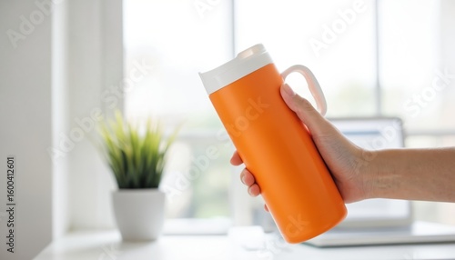Hand holds orange insulated stainless steel tumbler with handle. Perfect for hot or cold drinks like iced tea or coffee. Modern beverage container for travel, hydration, ideal for office or daily use.