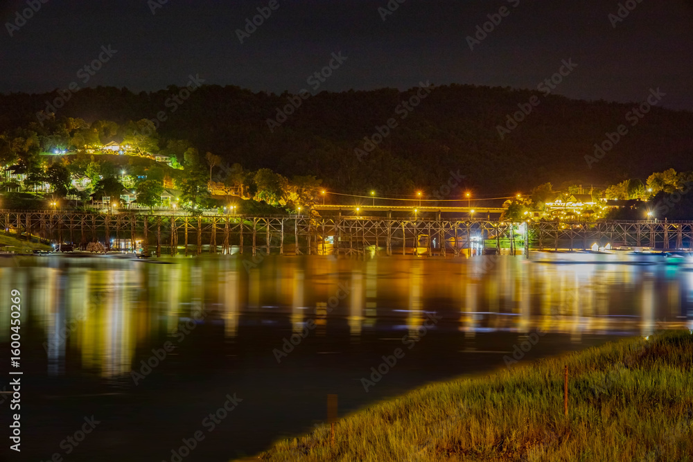Fototapeta premium The enchanting night view of Wooden Mon Bridge at Sangkla Buri Kanchanaburi, Thailand, this cityscape reflects city lights on the water with a bridge silhouetted against the evening sky.