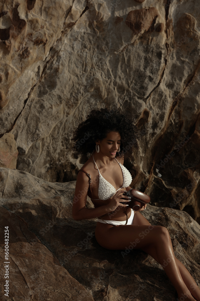 Naklejka premium A young woman with curly hair smiles, holding a vintage camera while sitting on sunlit rocks by the sea.