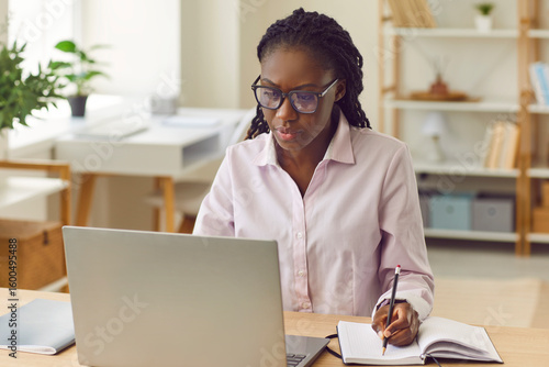 Fotografia Portrait of a concentrated serious african american business woman sitting in a shirt and glasses at the desk on her workplace and working on laptop at office looking at the monitor screen