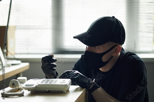 Foto Man wearing black cap and mask tampering with landline phone using screwdriver i