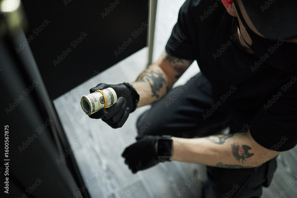 Naklejka premium Tattooed young man wearing black gloves kneeling on floor stealing roll of US dollar bills from broken safe, face partially visible, engaging in covert activity, during raid on corporate office