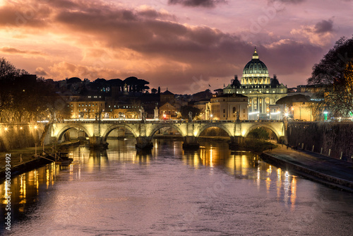 View of the illuminated Ponte Sant'Angelo gracefully spans the tranquil Tiber River towards the majestic St. Peter's Basilica at dusk, Rome, Lazio, Italy.