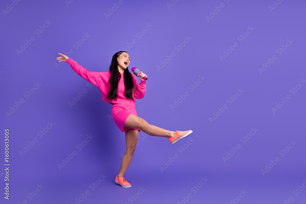 Cheerful young woman in pink outfit singing into a microphone while posed against a bright purple background Cheerful young woman in pink outfit singing into a microphone while posed against a bright purple background