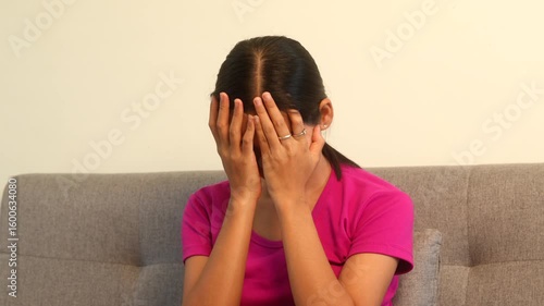 Asian woman sits on a sofa, feeling alone. Her expression reveals deep sadness, anxiety, and signs of depression or panic in a quiet room.