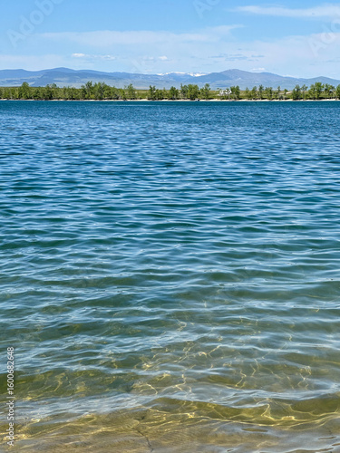 Vibrant blue lake water sparkles under the summer sun with a distant tree-lined shoreline and Montana mountains in the background.
