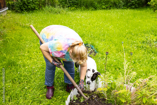 An older woman works in the garden, digging beds and watering. A black and white dog watches her work as a woman with a shovel