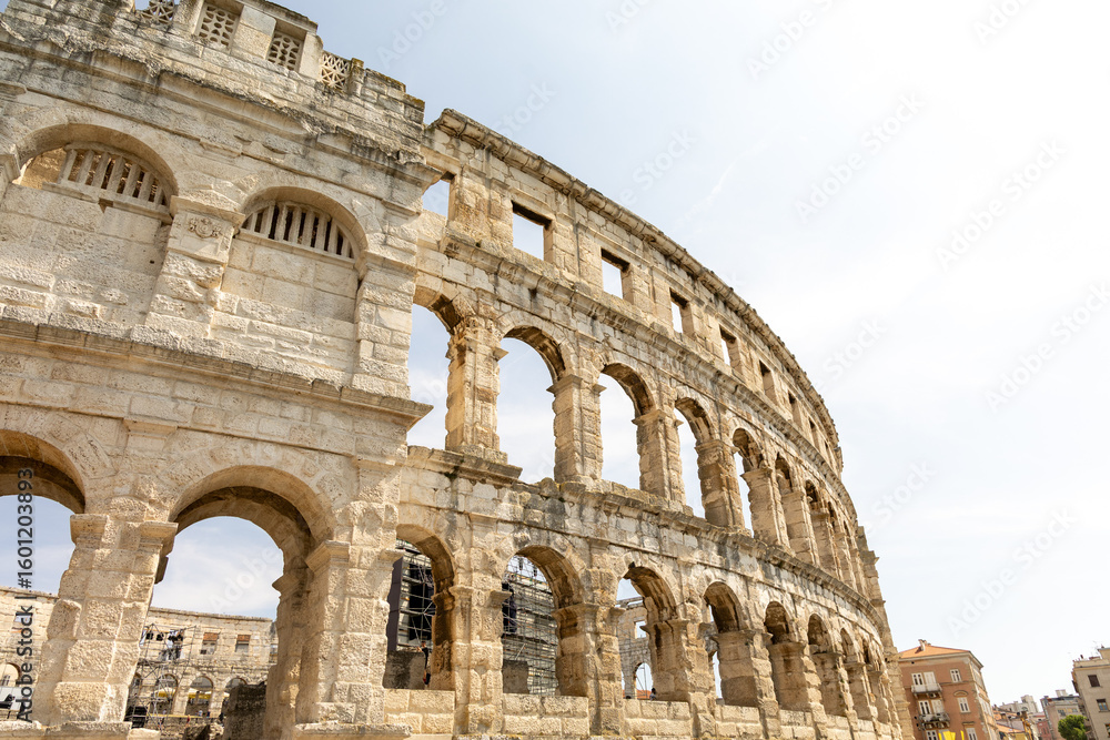 Naklejka premium Ancient amphitheater showcasing impressive stone architecture, arches, and historical significance, surrounded by modern urban landscape and clear sky
