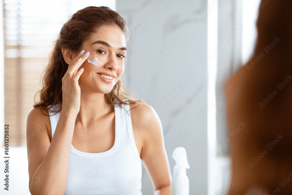 Fototapeta premium Happy Woman Applying Face Cream Moisturizing And Caring For Skin Smiling To Her Reflection In Mirror Standing In Modern Bathroom At Home. Facial Skincare And Pampering. Selective Focus