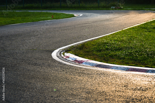 Smooth asphalt racing track making a soft curve, with red-and-white curb markings and lush green grass alongside. Captured during golden hour, the warm light enhances the texture of the pavement