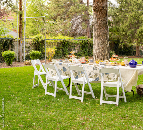 An outdoor tea party table set up in a garden with white folding chairs, floral tablecloth, snacks, tea sets, and tiered trays