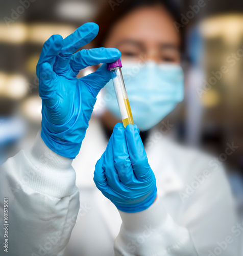 Close up of a lab technician's gloved hands holding a test tube filled with yellow liquid. The technician is female and her face is diffused in the background