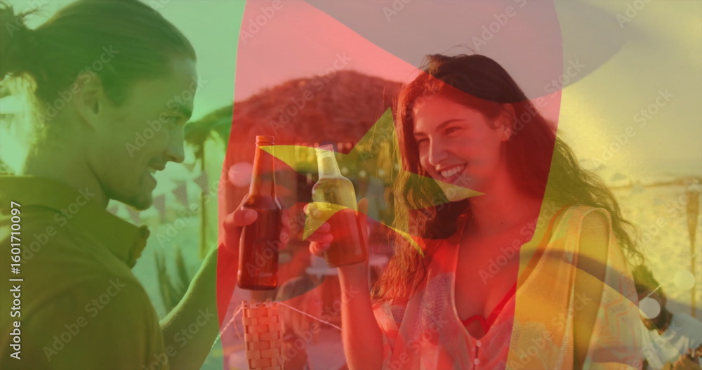 Fototapeta premium Raising casually dressed couple toasting beer bottles under straw umbrellas at beach lounge