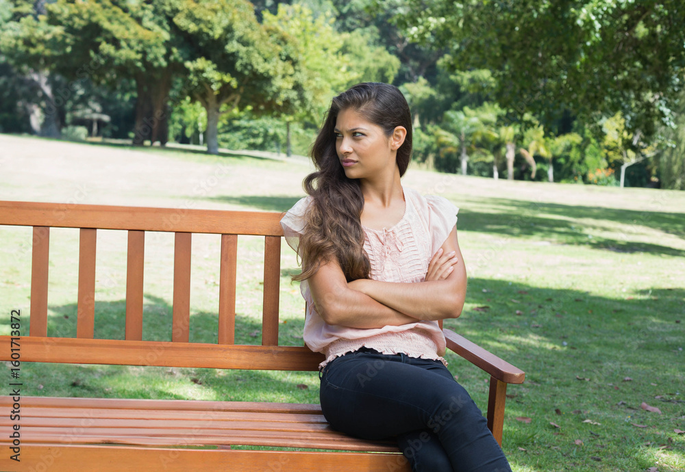 Obraz premium Woman sitting on wooden slatted bench in green park, crossing arms and looking left contemplatively