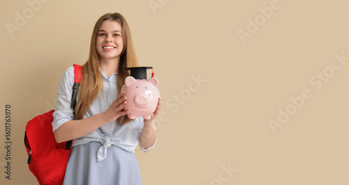 Female student with piggy bank in graduation hat and backpack on beige backgr...