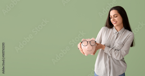 Beautiful young happy woman with piggy bank in eyeglasses on green background...