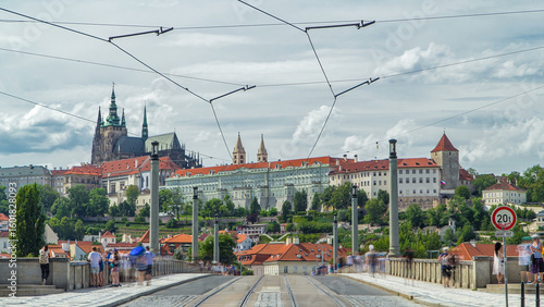 Photography Red tram passing on Manes Bridge timelapse and famous Prague Castle on the background in Prague, Czech Republic