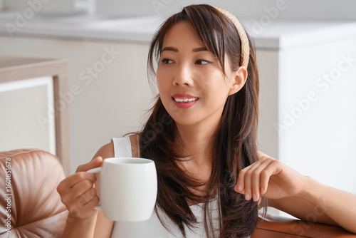 Young Asian woman with cup of coffee sitting on sofa in kitchen, closeup